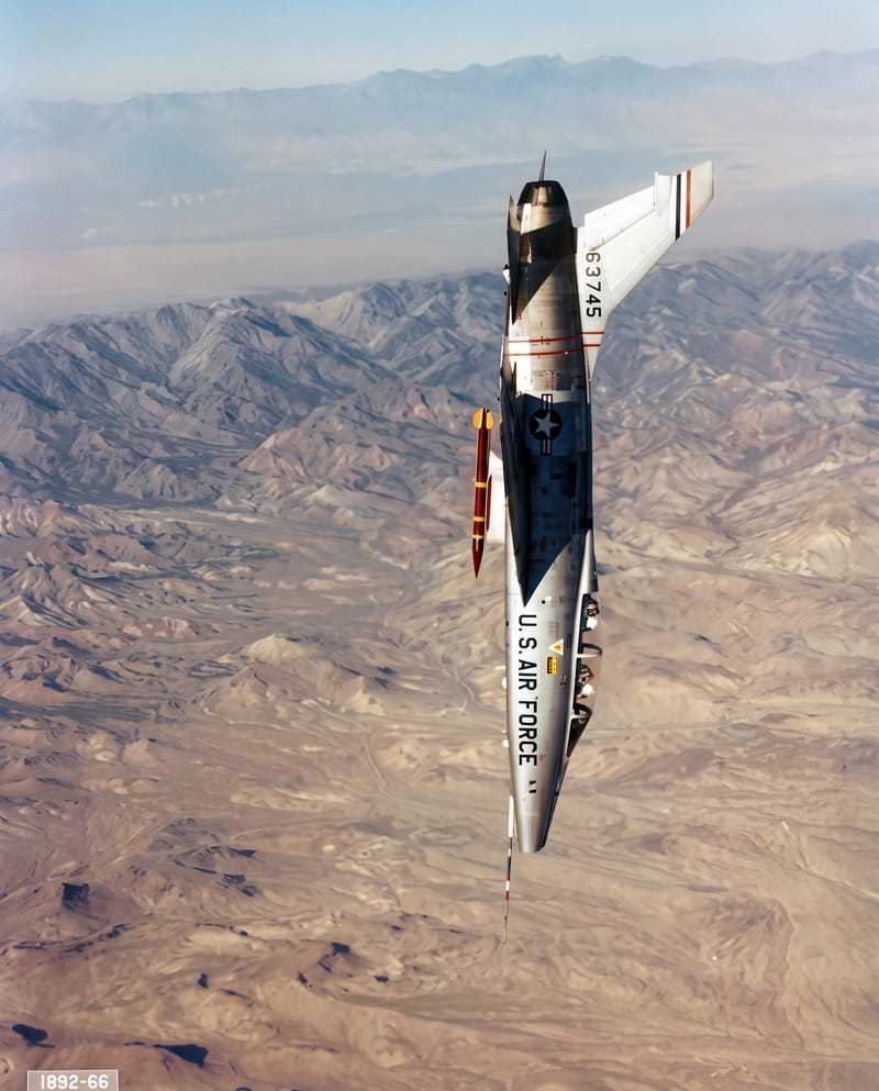 A silver U.S. Air Force F-100 jet in a steep, nose-down dive above brown desert mountains.