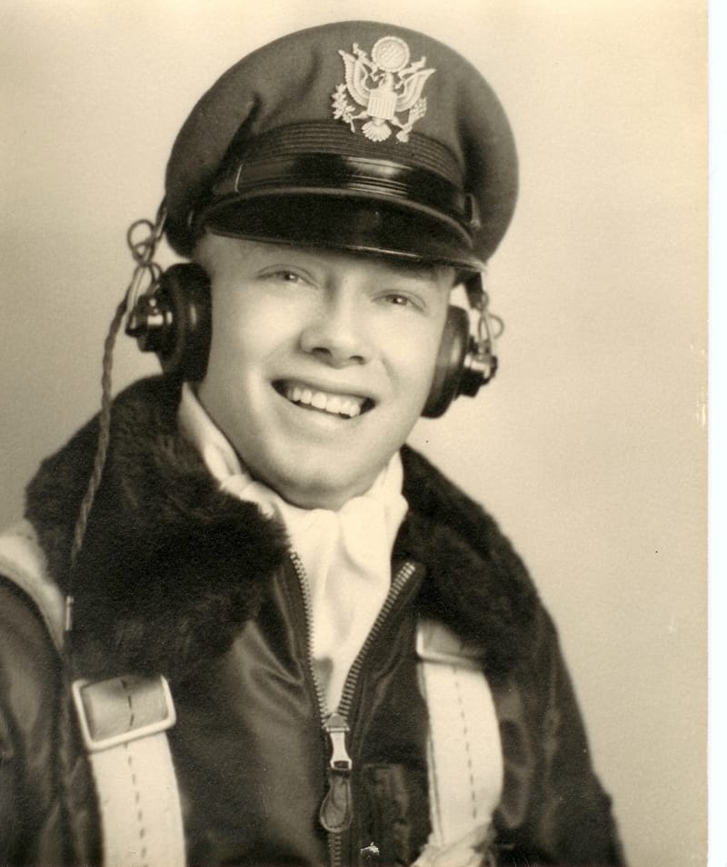 Black and white portrait of a young, smiling airman wearing an officer’s cap, a headset, and a fur-collared flight jacket.