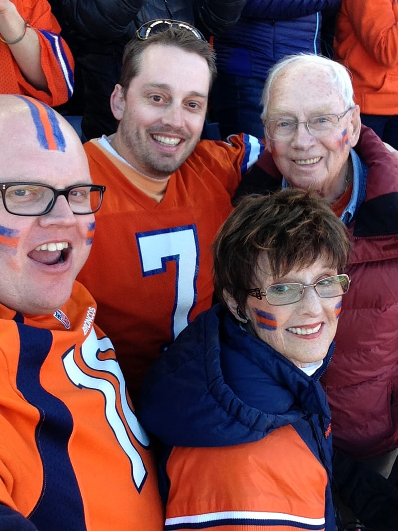 Four people in orange and navy Denver Broncos gear with team colors painted on their faces, crowded together and smiling for a selfie.