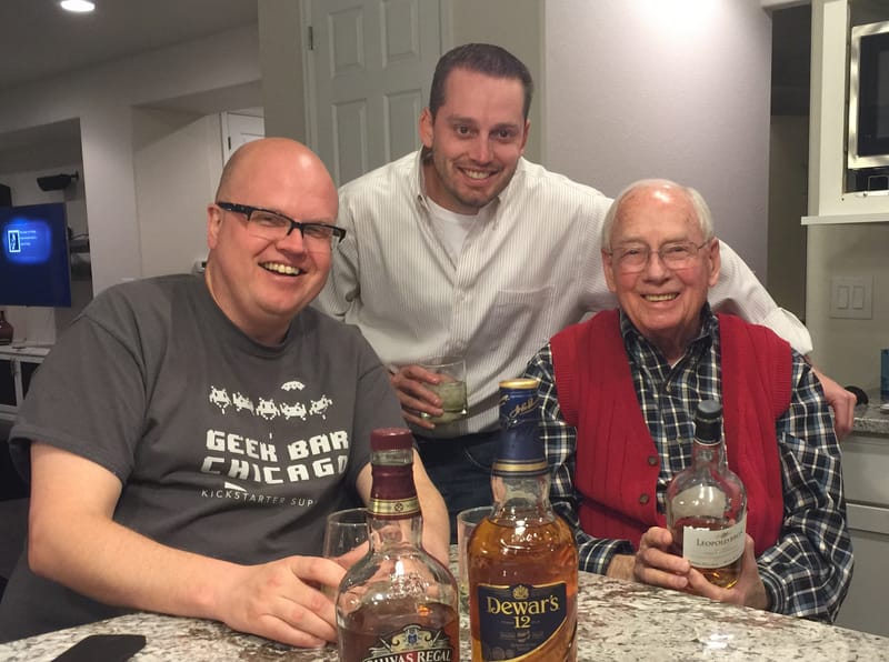 Don in a red vest, seated at a kitchen counter with two smiling younger men and several bottles of whisky.