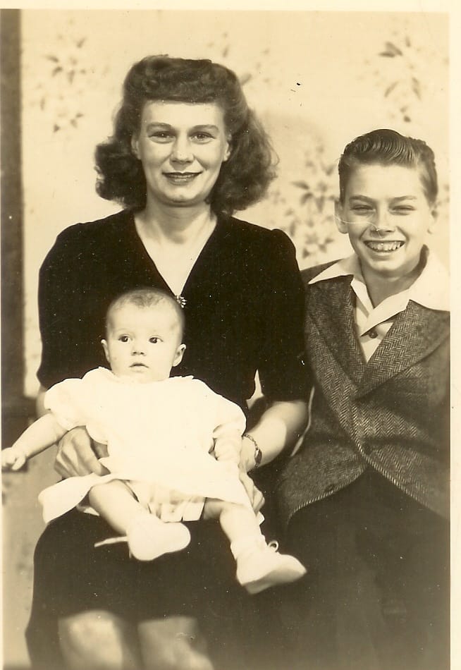 Sepia portrait of a smiling woman holding a baby in a white dress, with a grinning boy in a jacket beside her.