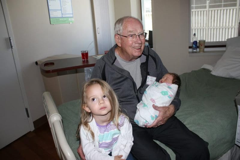 Don sitting on a hospital bed and beaming, holding a swaddled newborn, with a young blond girl standing in front of him.