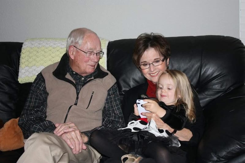 Don in a tan vest on a black leather couch, smiling beside a woman in glasses and a young blond girl who is holding a phone.