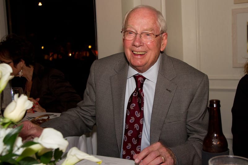 An older man with glasses, a gray jacket, and a paisley tie, laughing at a dinner table with white flowers.