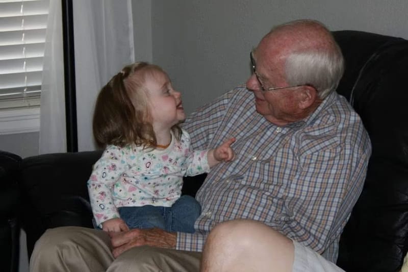 Don on a black leather couch in a plaid shirt, with a toddler on his lap pointing at him and looking up at his face.