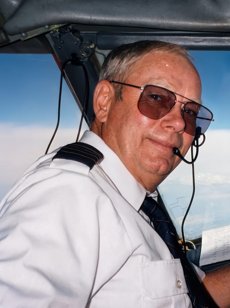 A smiling pilot in a white uniform shirt, tinted aviator glasses, and a headset, looking back from the cockpit with blue sky behind him.