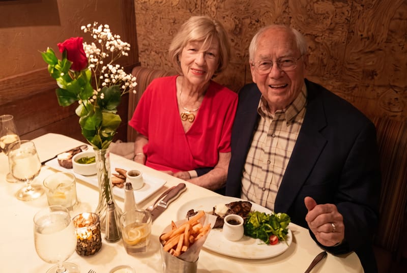 Don and Woodie sitting close together at a restaurant table with a single red rose. She wears a red dress and he gives a thumbs up.