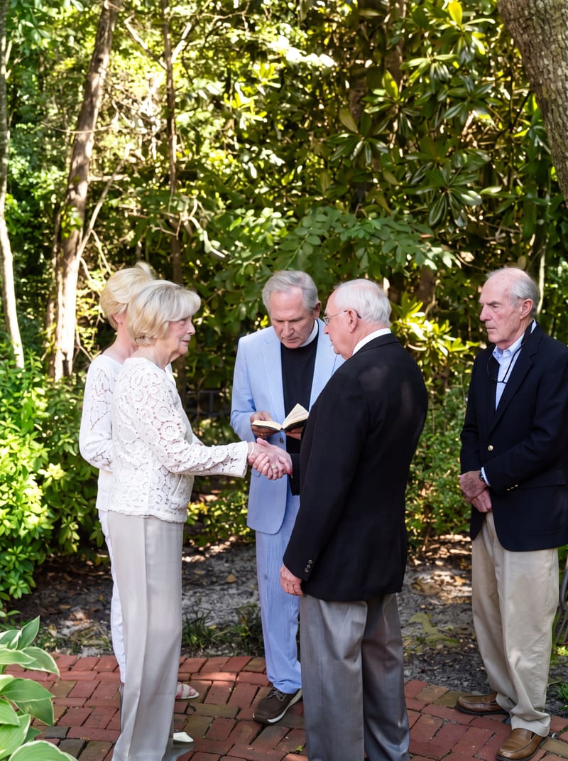 Don and Woodie holding hands before an officiant in a light blue jacket during an outdoor ceremony among green trees, with two guests beside them.